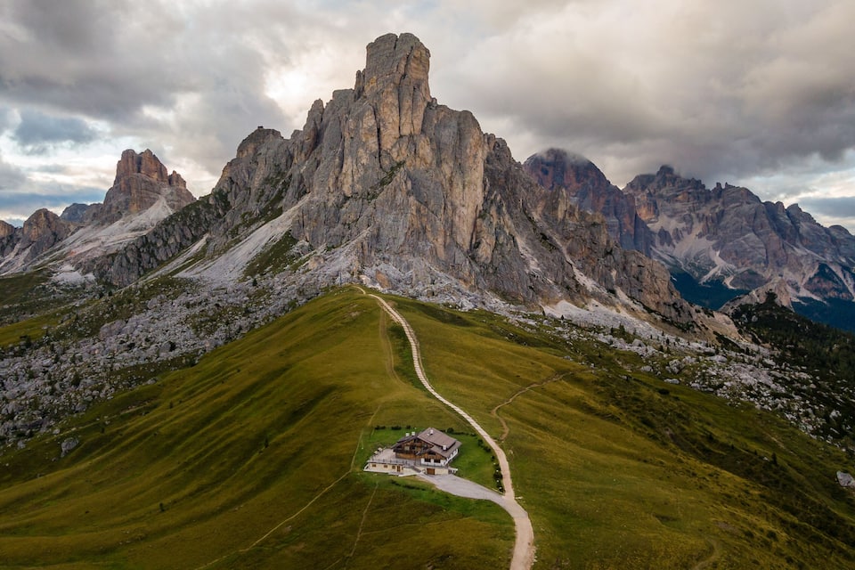 A mountain landscape with a prominent rocky peak, a winding path leading to a small building, and surrounding green hills.