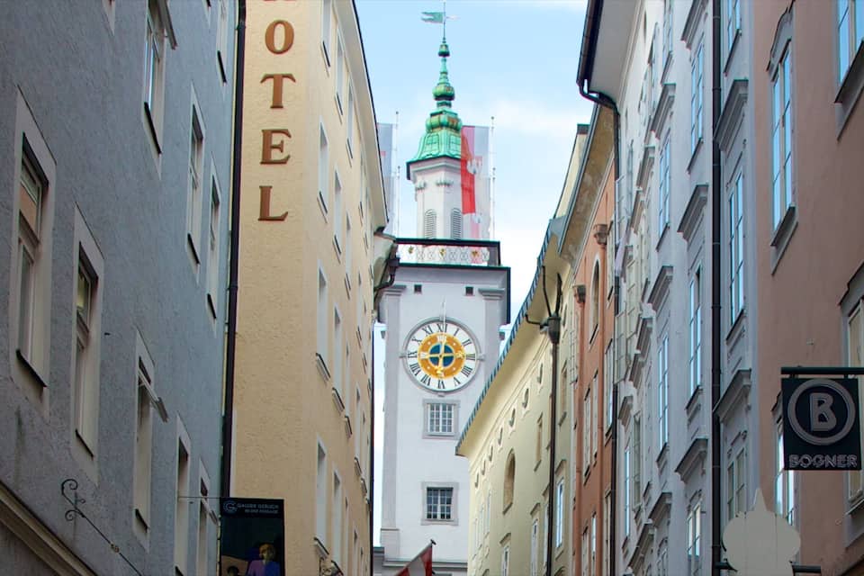 A narrow street with a clock tower, a hotel, and a building with a 'B' sign.