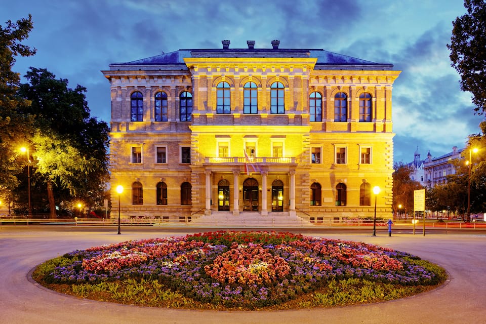 Un edifici històric amb una gran entrada central i un jardí de flors al davant.
