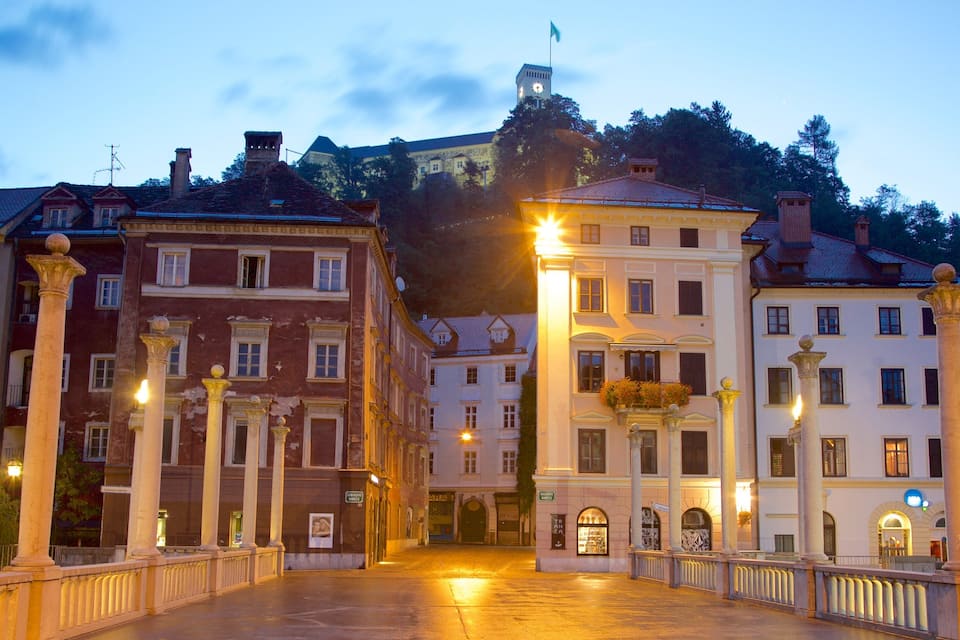 A historic town square with classical architecture, a prominent clock tower, and a mountain backdrop.
