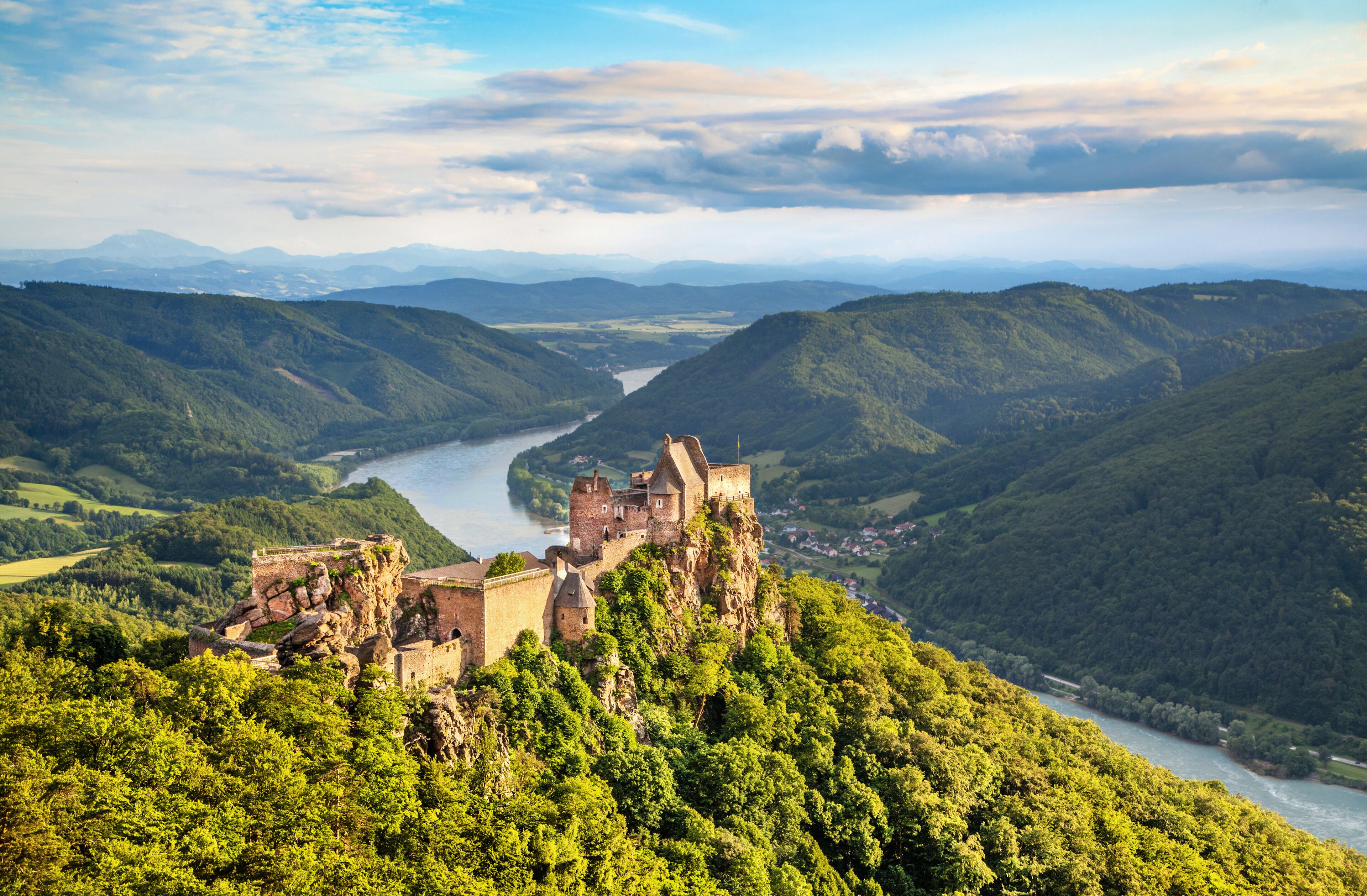 A historic castle perched on a cliff overlooking a river valley with lush greenery and distant mountains.
