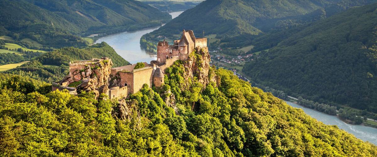 A historic castle perched on a cliff overlooking a river valley with lush greenery and distant mountains.