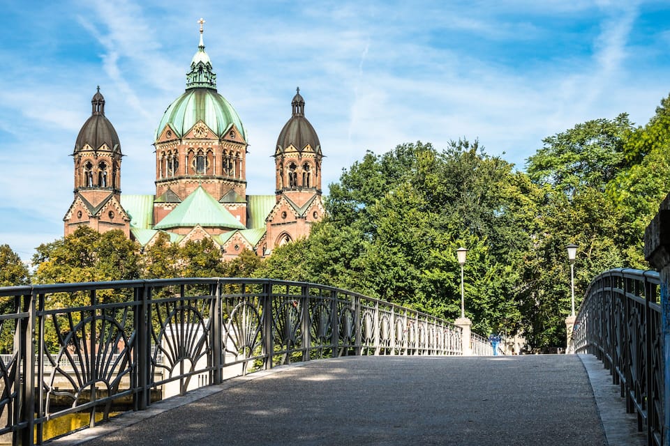 A bridge with ornate metal railings, leading to a large church with three domes and a green roof.