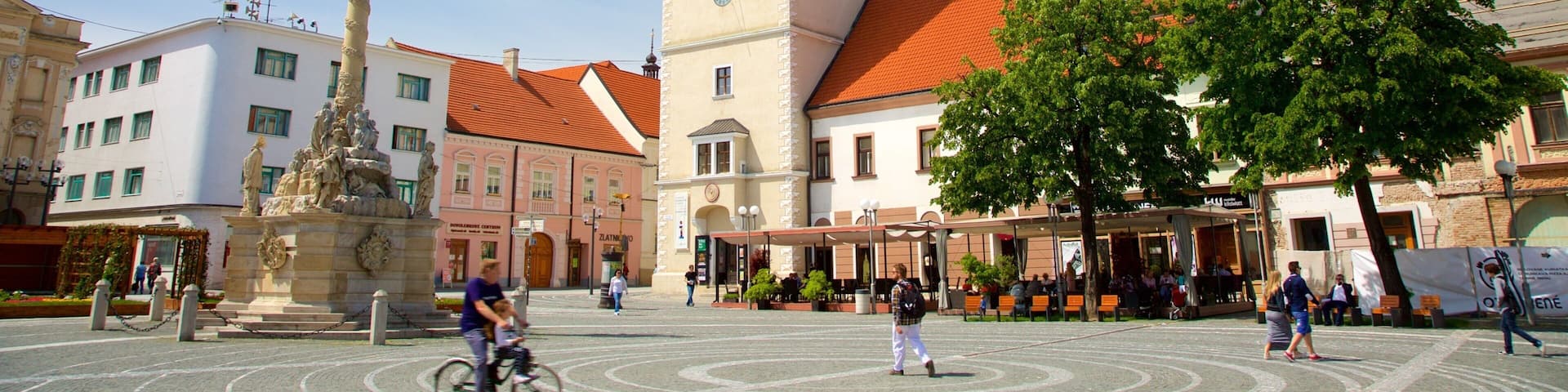 A square with a clock tower, a statue, and surrounding buildings.
