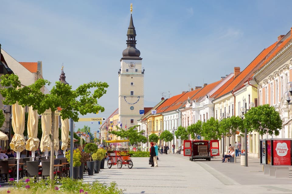 A European city street with historic buildings, a prominent clock tower, and outdoor seating areas.