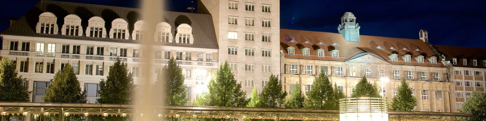 Un edificio histórico con una torre de reloj, una fuente y una plaza bien iluminada con árboles y tiendas.