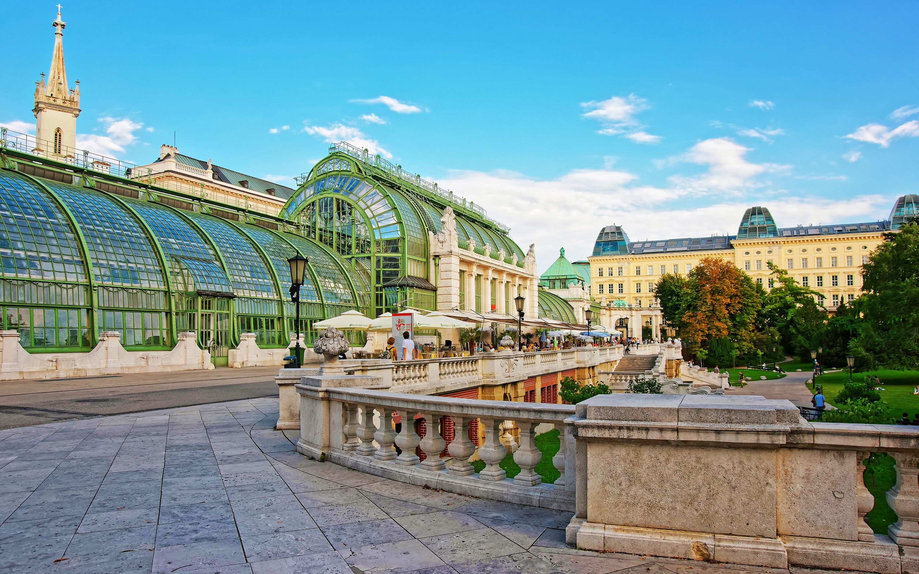 Ein historisches Gebäude mit Glasdach und einem markanten Turm, eine Brücke mit steinernen Geländern und ein gepflegter Gartenbereich.