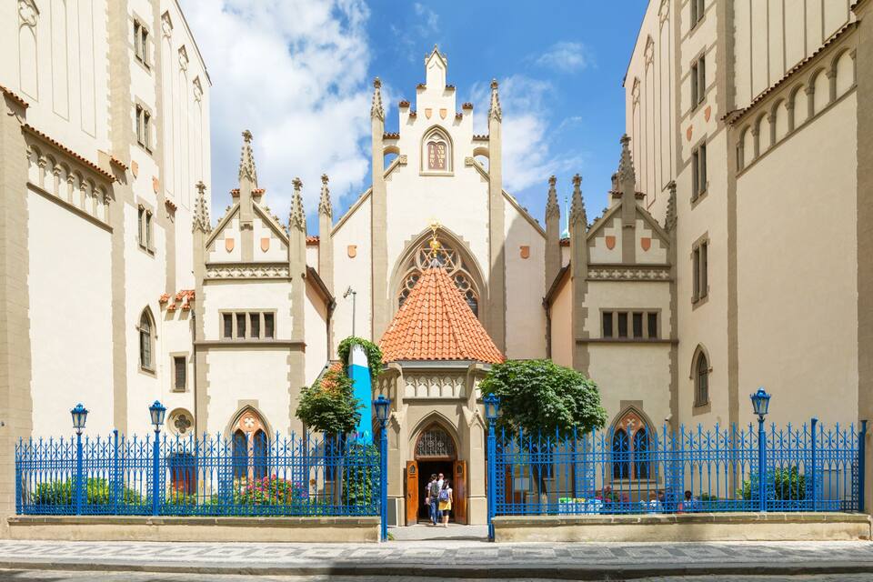 A historic building with a red-tiled roof, ornate Gothic windows, and a blue iron fence.