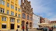 A historic European street with yellow and red brick buildings, including a pharmacy and a restaurant.