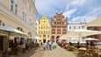 A cobblestone square with outdoor cafes, historic buildings, and people walking.