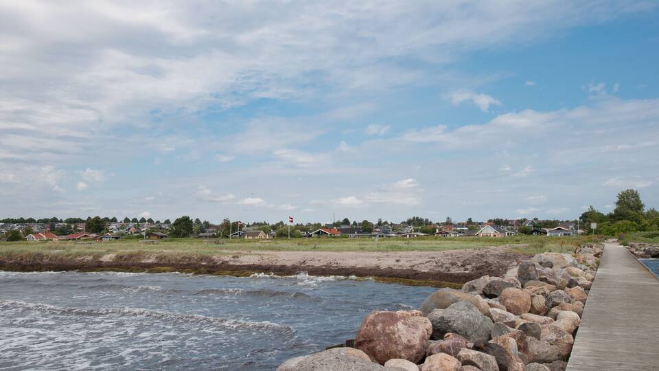 A wooden pier extending into a body of water, surrounded by large rocks and a distant shoreline with houses and a flag.