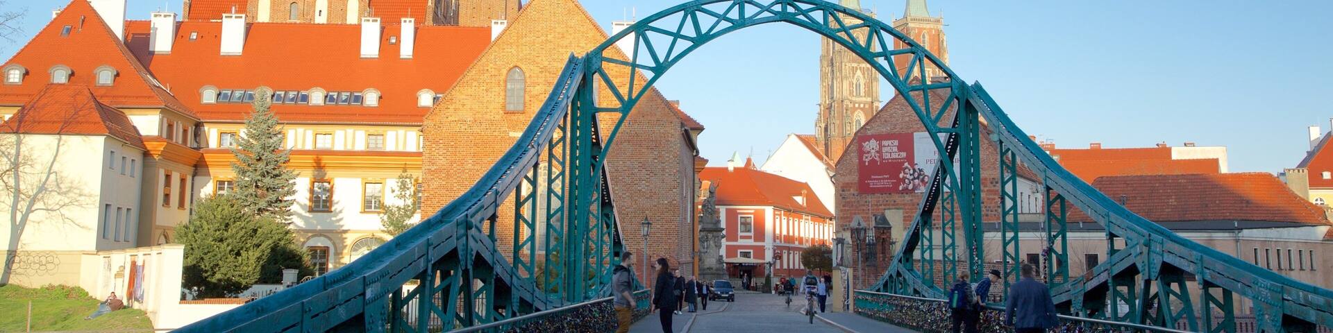 Un puente histórico con un arco de metal verde, que lleva a una ciudad con edificios de techo rojo y una iglesia destacada con dos torres.