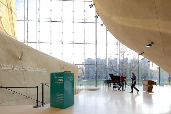 Un bâtiment moderne doté de grandes fenêtres et d’un plafond incurvé. On y voit une enseigne verte avec du texte blanc, un piano et une personne qui marche.