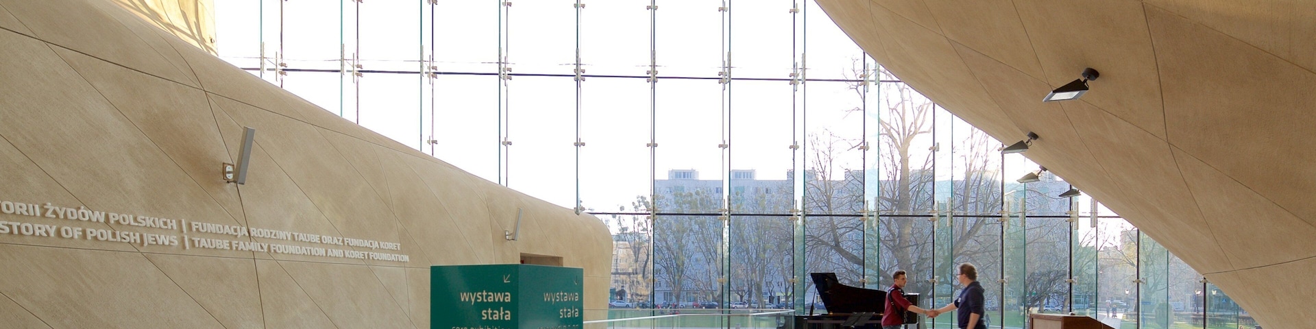 Un edificio moderno con amplios ventanales y techo curvo. Se observa una señal verde con letras blancas, un piano y una persona caminando.