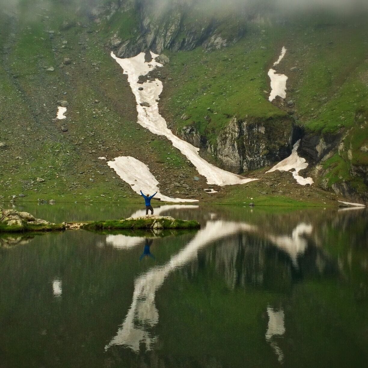 Una persona parada en una pequeña isla en un lago, con parches de nieve en las montañas circundantes.
