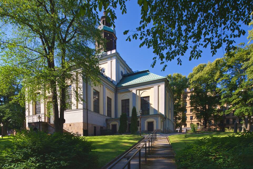 A historic building with a green roof, surrounded by lush greenery and a clear blue sky.