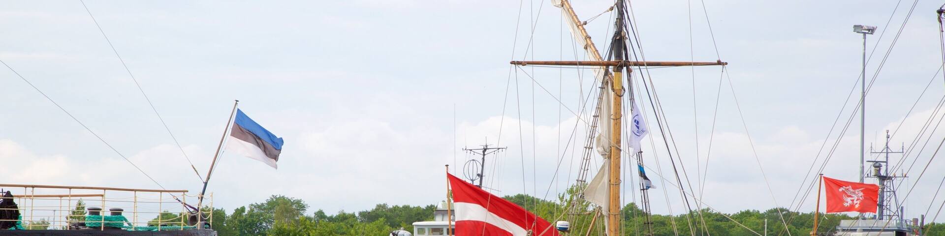 A historic sailing ship docked at a harbor with other vessels and a flag.