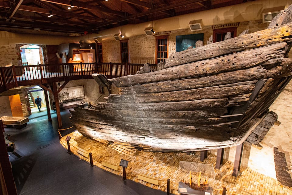 A large, weathered shipwreck displayed indoors, surrounded by stone walls and wooden beams.