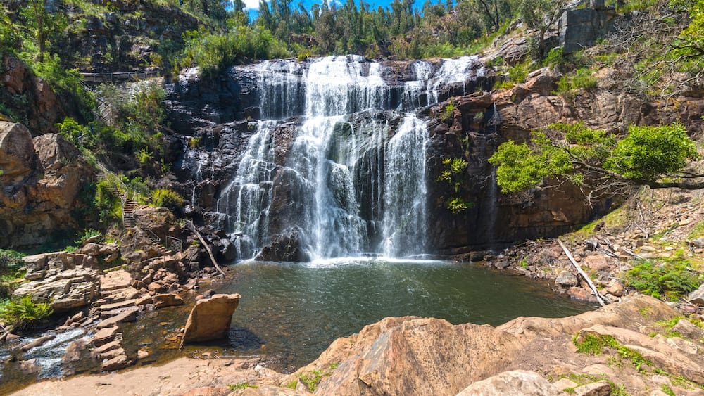 Ein mehrstufiger Wasserfall stürzt in ein Becken, umgeben von felsigem Gelände und vereinzelter Vegetation.