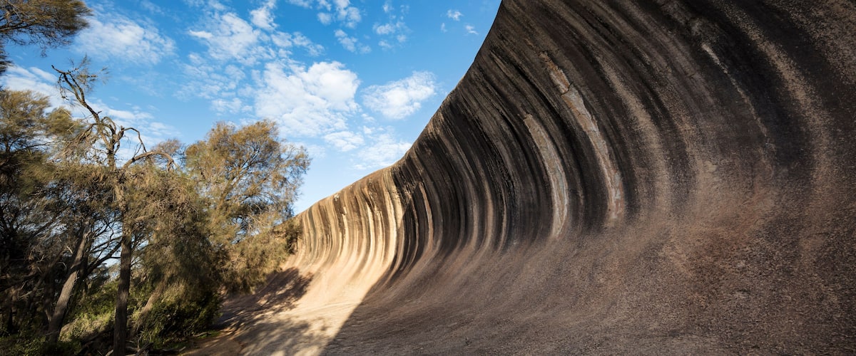 A large rock formation with a curved overhang, surrounded by trees and a clear blue sky.