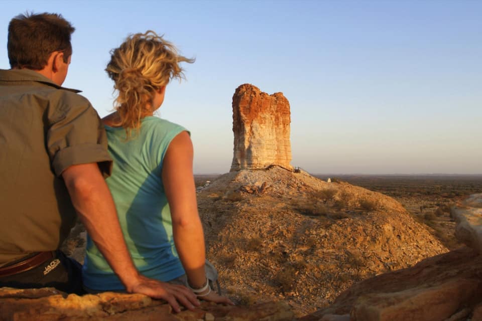 Un couple est assis sur un promontoire rocheux, d’où il domine une haute formation rocheuse érodée au milieu d’un paysage désertique.