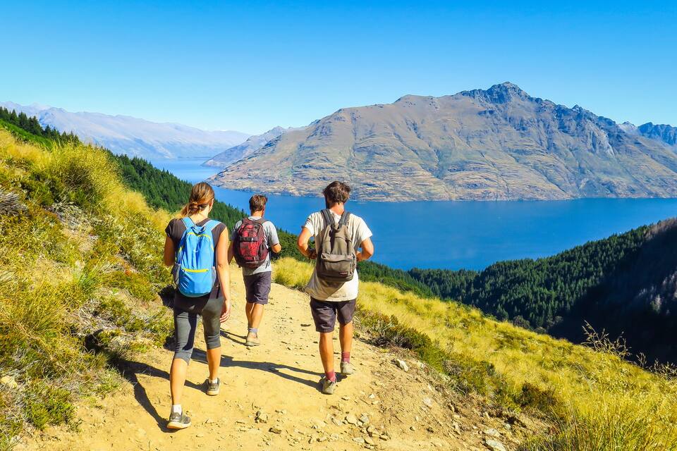 Hikers on a dirt trail with a mountainous backdrop and a lake.