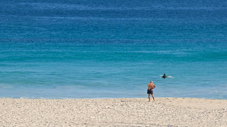 Une plage où une personne est debout et une autre fait de la planche à voile dans l’eau.