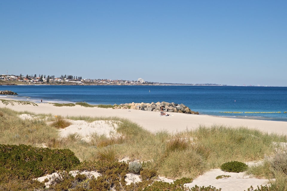 Platja sorrenca amb dunes i vegetació escassa, oceà blau i translúcid, i un poble allunyat amb edificis.