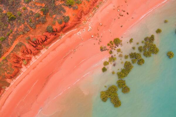 An aerial view of a beach with a distinct red sand area, clear water, and scattered green vegetation.
