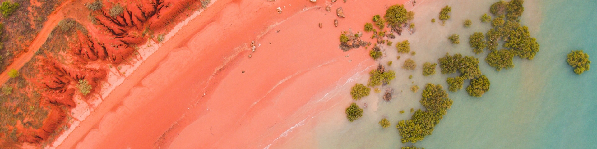 An aerial view of a beach with a distinct red sand area, clear water, and scattered green vegetation.