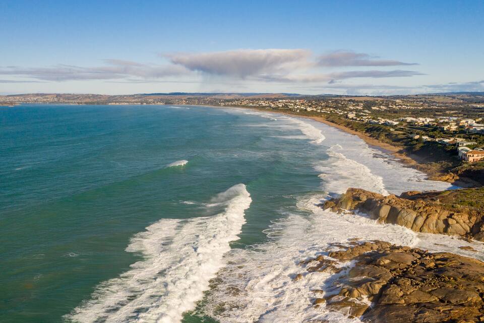 A coastal landscape with a rocky shoreline, waves crashing onto the beach, and a distant town with buildings.