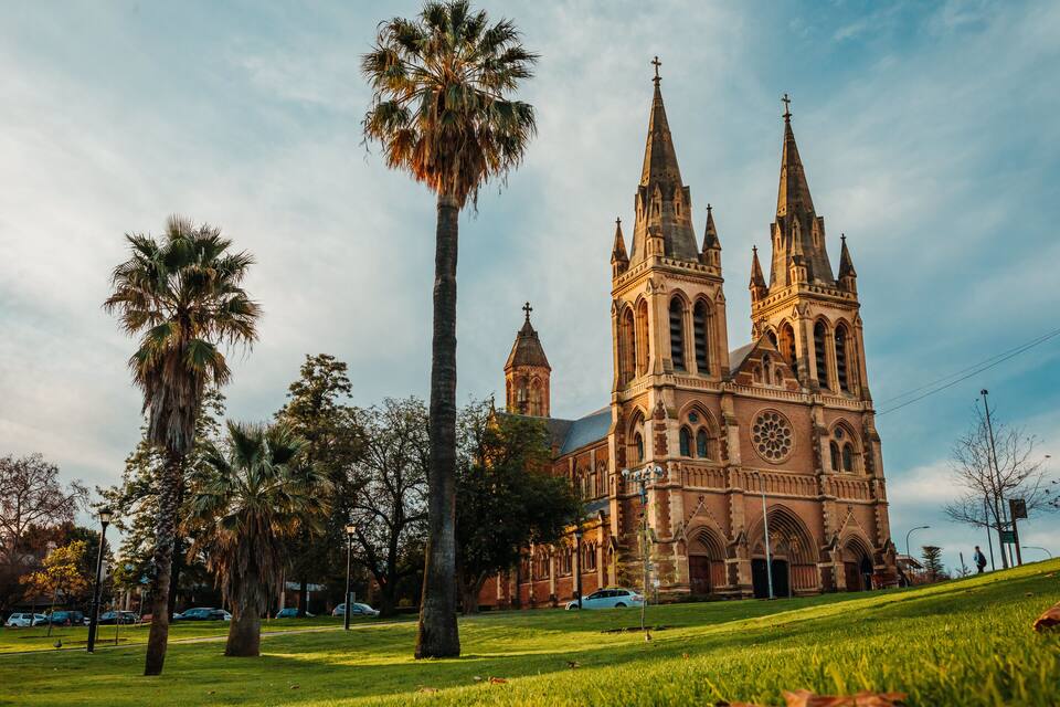A historic church with multiple spires and a large clock tower, surrounded by palm trees and a grassy area.