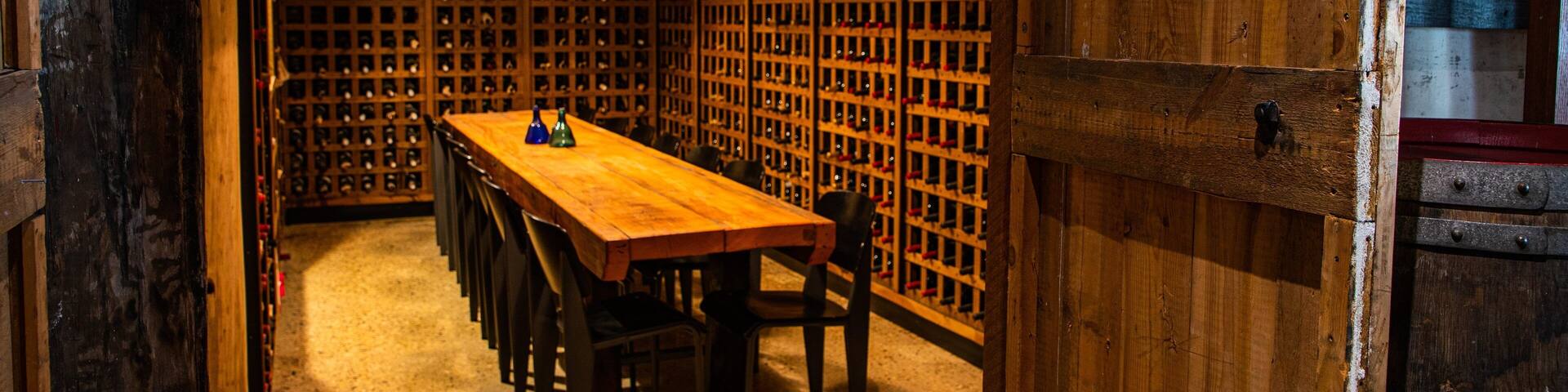 A wooden wine cellar with a long table and chairs, and a wall of wine racks.
