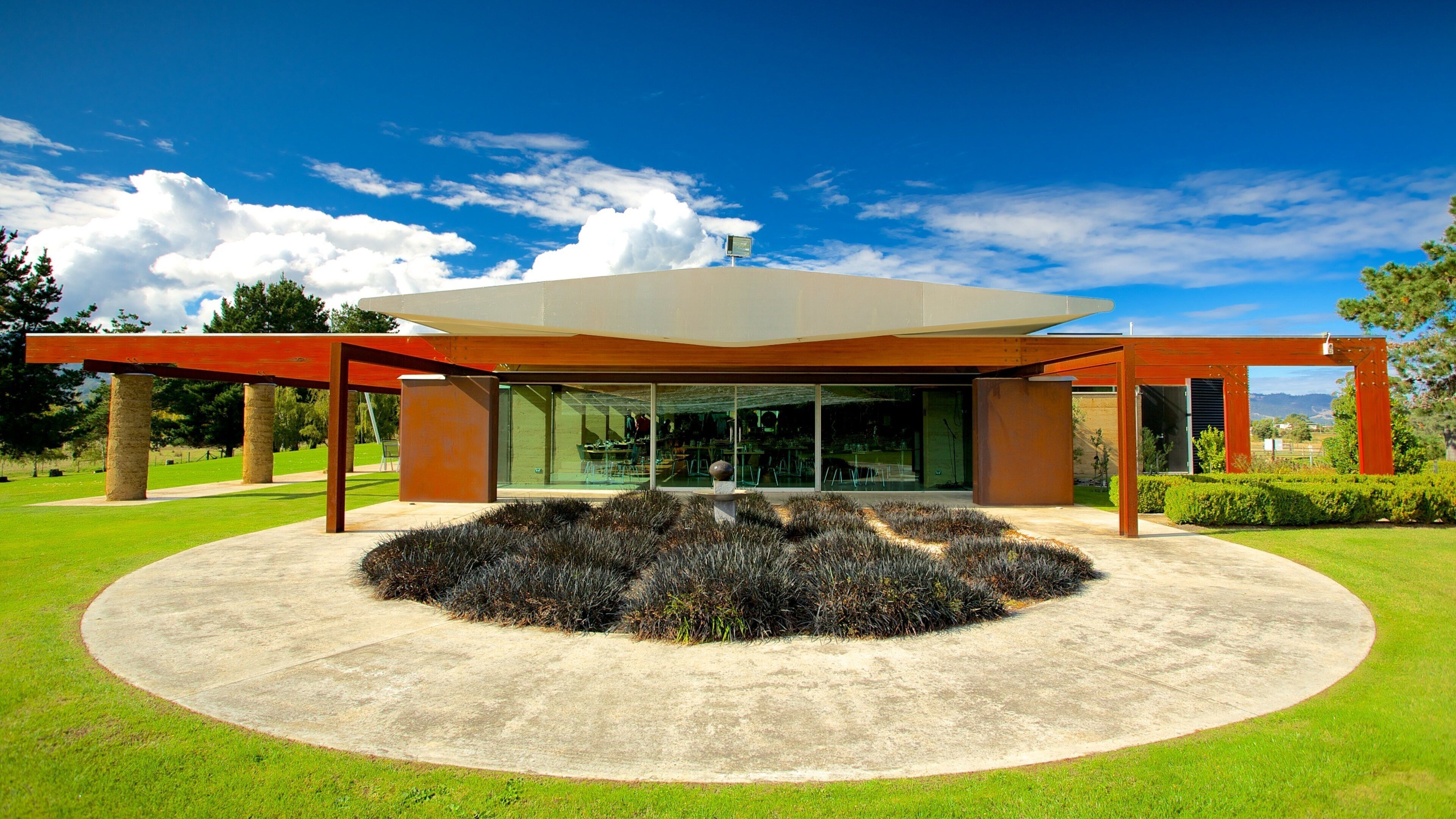 A modern building with a glass facade, surrounded by a circular garden and red beams.