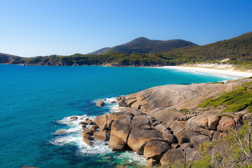 A coastal landscape with a rocky shoreline, clear blue sea, and a distant beach.