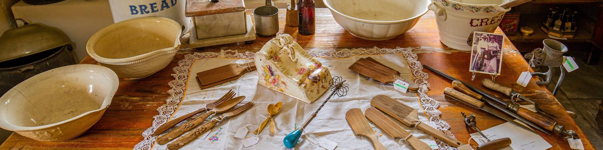 Una cocina vintage con utensilios de cocina antiguos, incluyendo un recipiente para pan, un tazón grande y diversas herramientas sobre una mesa con encaje.