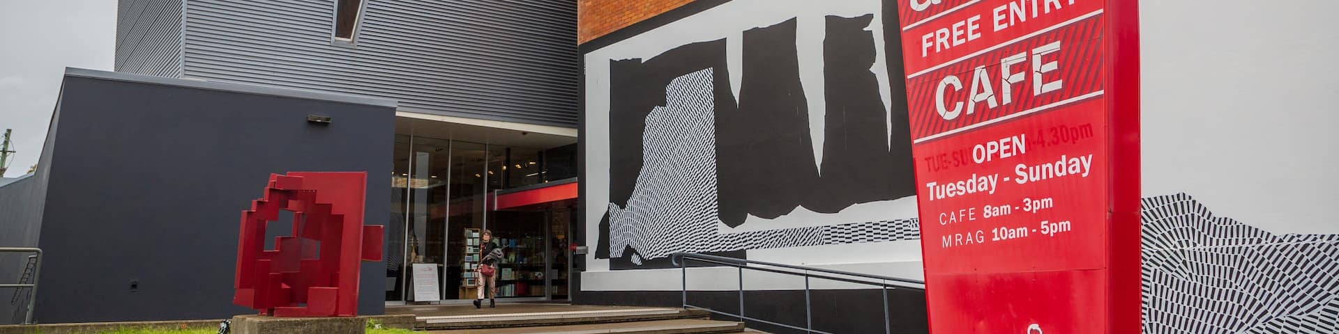 Un bâtiment moderne orné d’une grande fresque murale, d’une sculpture rouge et d’une enseigne indiquant la présence d’une galerie d’art et d’un café.