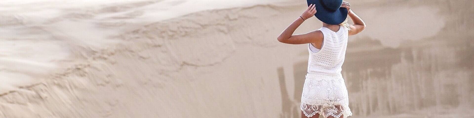 A person in a white dress standing on a sandy beach with a large sand dune in the background.