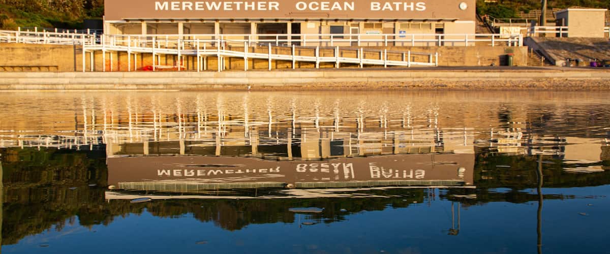 Een gebouw met de tekst 'Merewether Ocean Baths' en een weerspiegeling in het water.