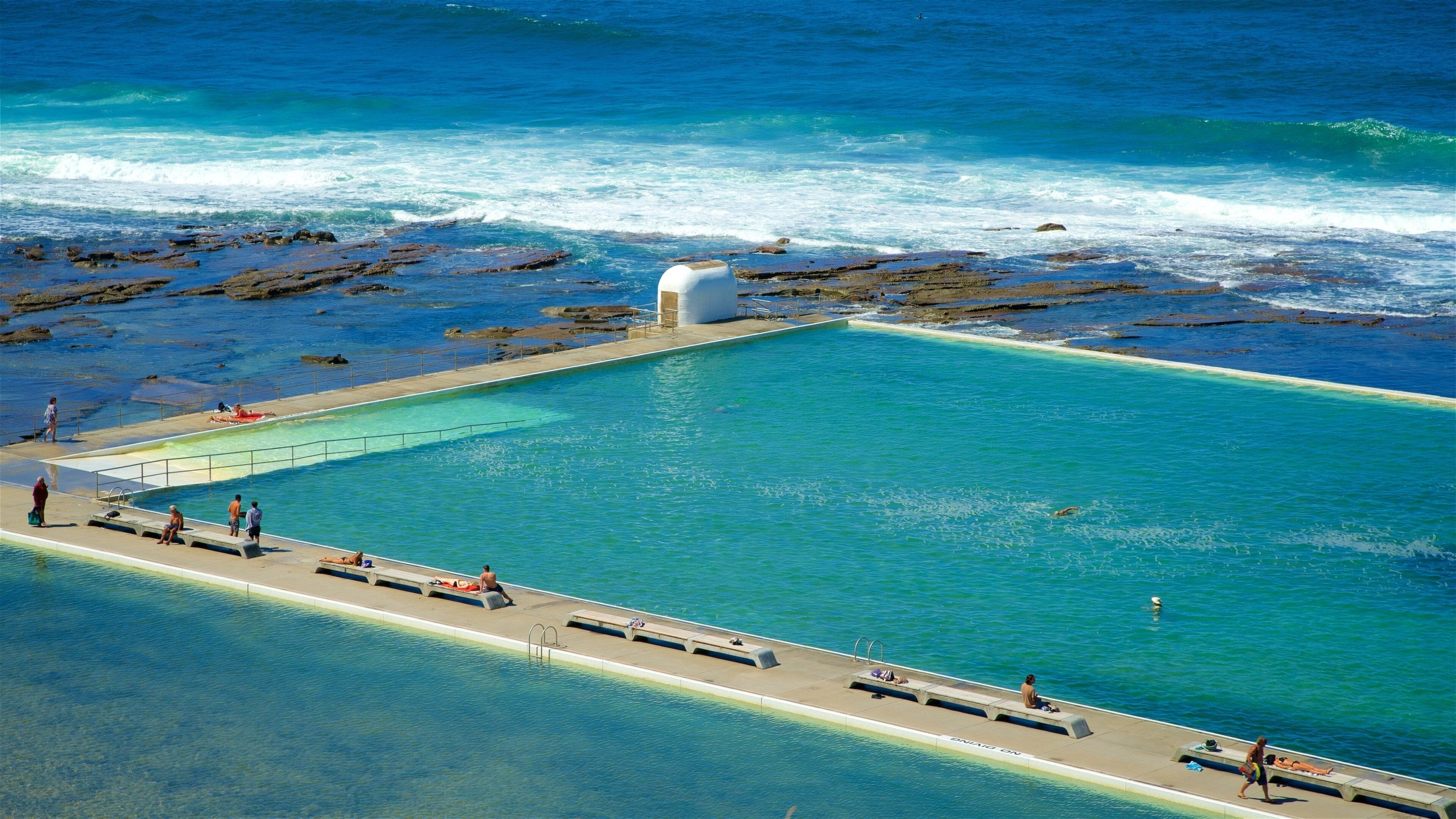 A large, clear blue pool with a concrete deck, surrounded by rocky terrain and the ocean.
