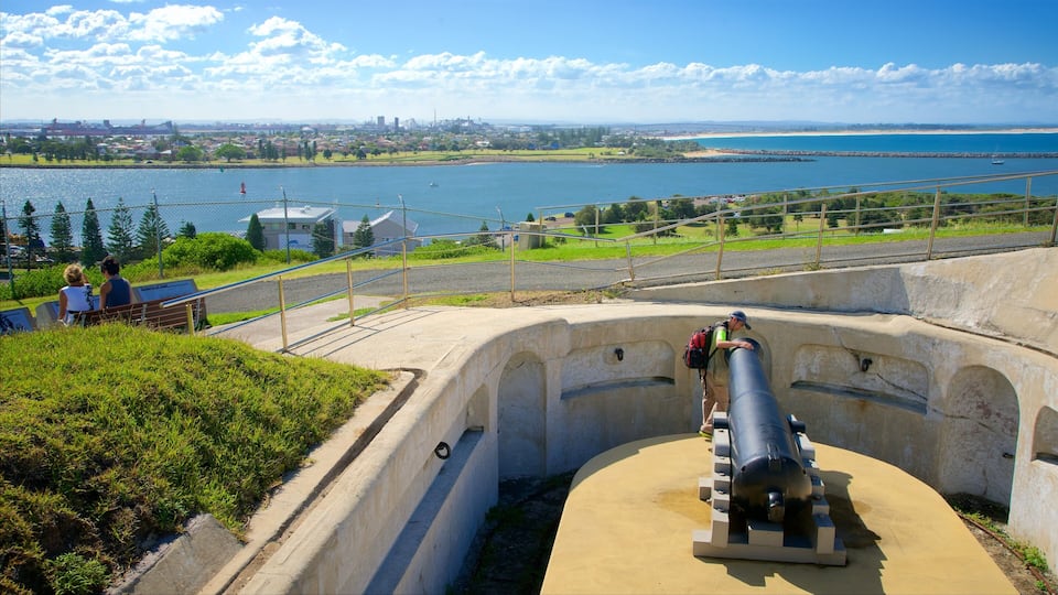 Um canhão em uma fortificação de concreto com vista para um corpo d'água e um horizonte urbano ao fundo.