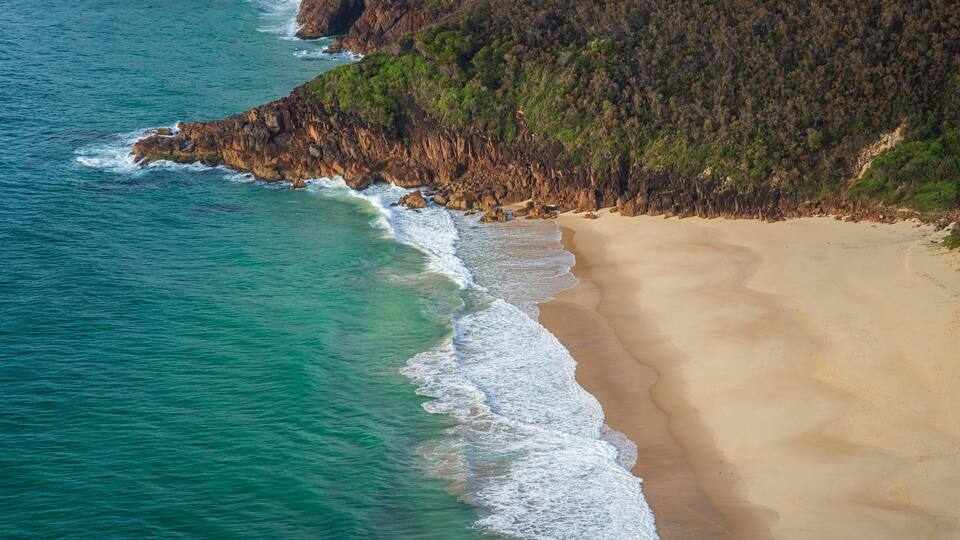 A coastal landscape with a sandy beach, turquoise ocean waves, and a forested cliff.