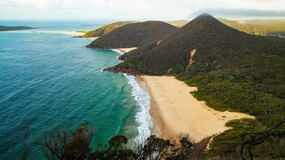 A coastal landscape with a sandy beach, turquoise waters, and forested hills.