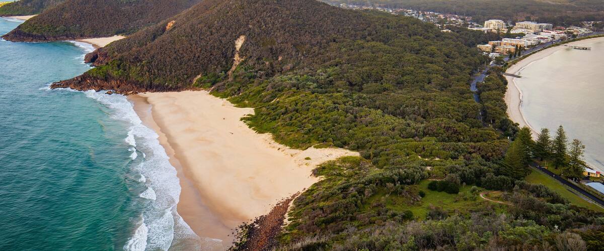 A coastal landscape with a beach, forested hills, and a town in the background.