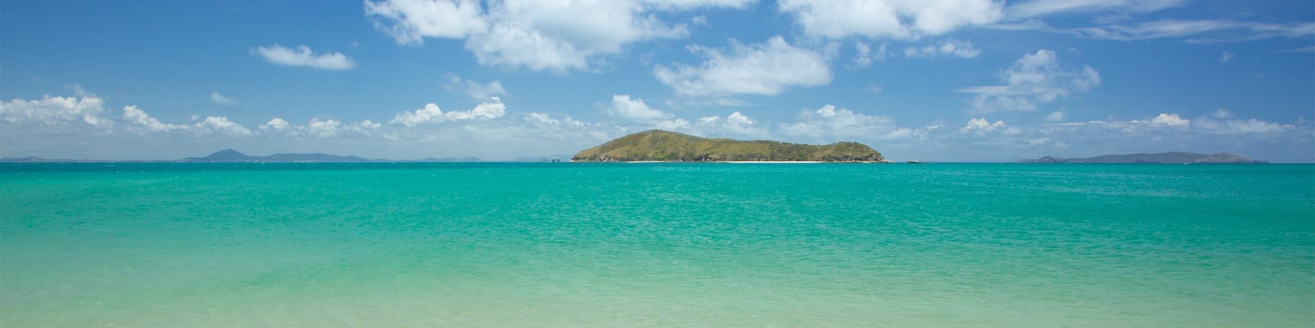 Una playa con agua turquesa cristalina, una orilla arenosa y una pequeña isla a lo lejos.