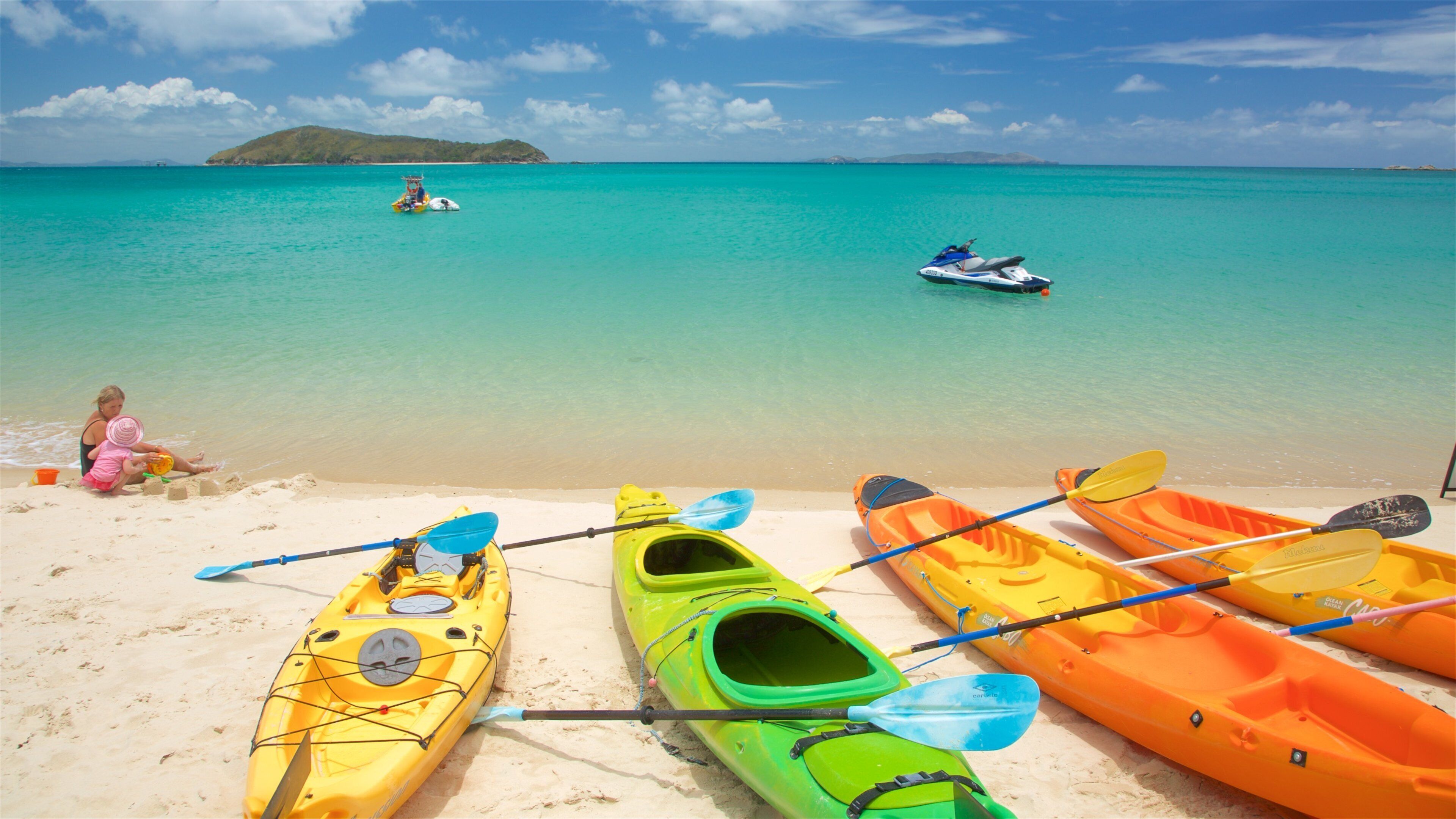 A beach with kayaks and a jet ski, a woman and child playing with sand, and a clear blue sea with a distant island.
