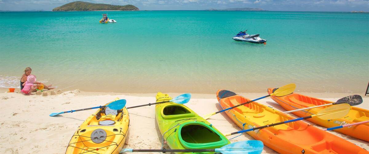 A beach with kayaks and a jet ski, a woman and child playing with sand, and a clear blue sea with a distant island.