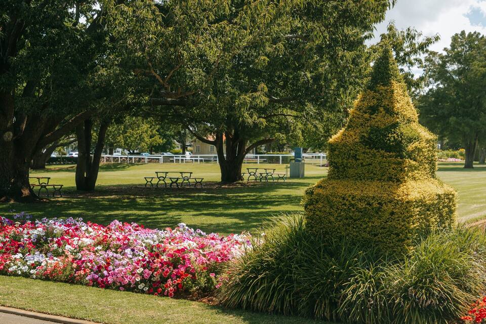 A park with a topiary tree, picnic tables, and flower beds.