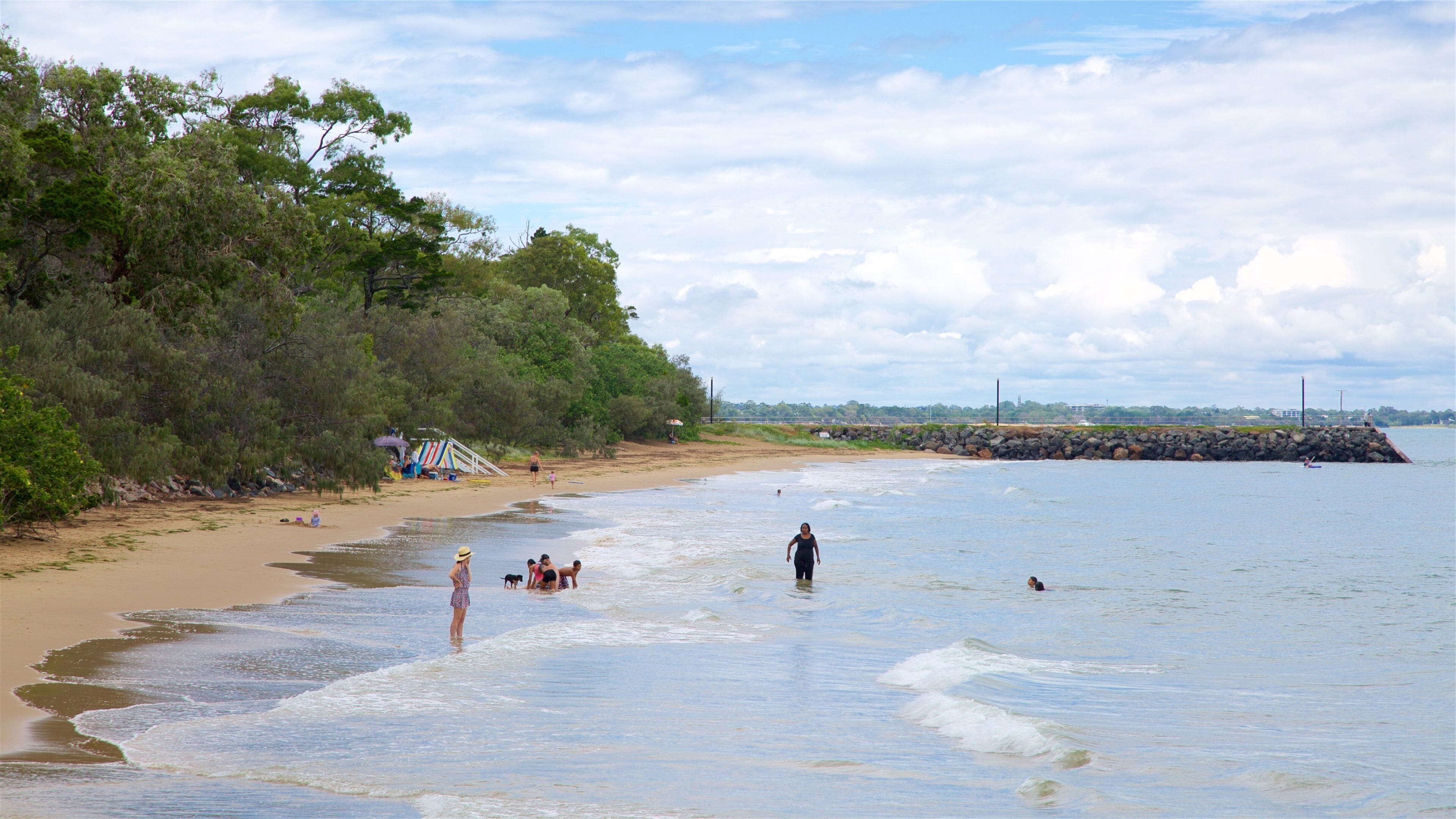 A beach with people swimming and walking, a rocky breakwater, and dense trees on the shore.