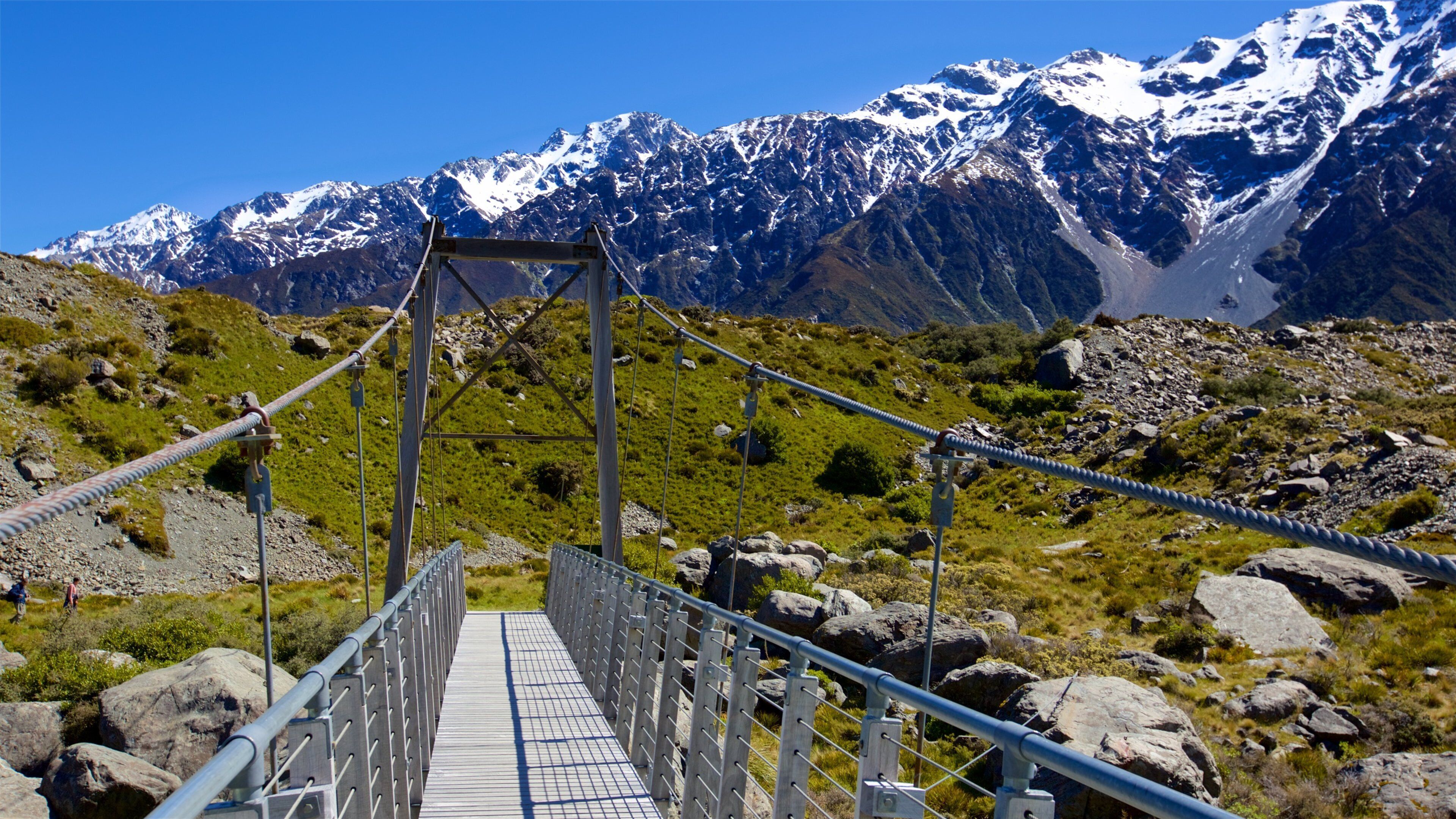 A suspension bridge with a metal railing, surrounded by rocky terrain and snow-capped mountains in the background.